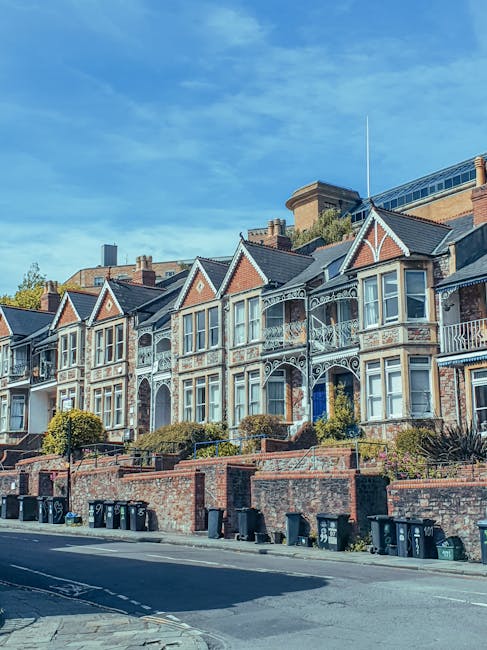 Photograph of a residential neighborhood featuring red-brick terraced houses with tiled roofs and white-framed windows. The houses are aligned along a paved walkway that curves through the area, borde