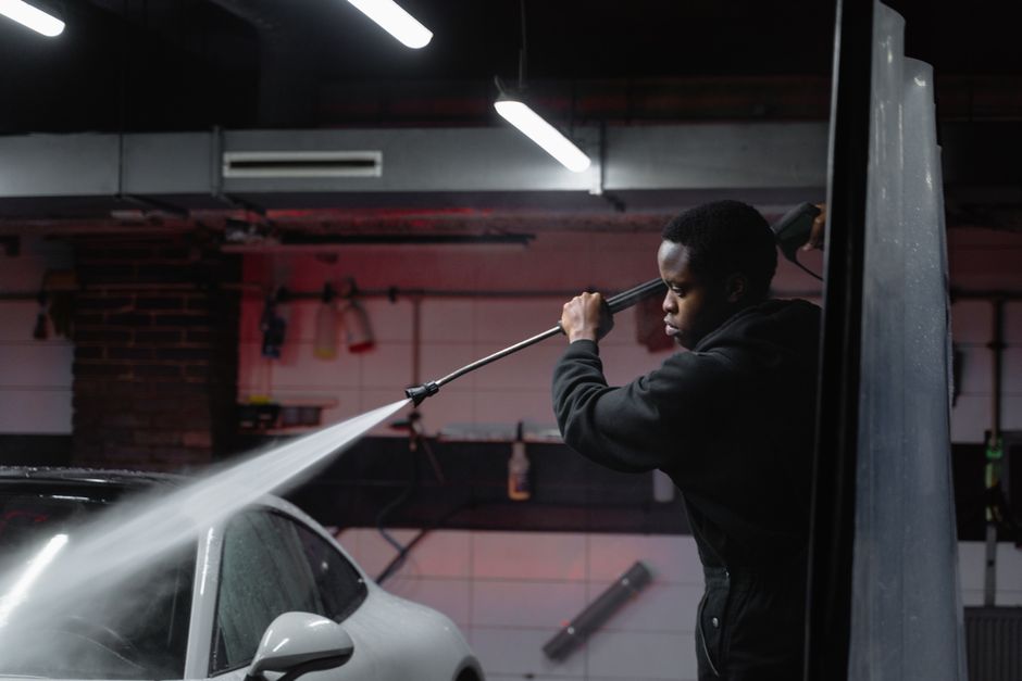 A person wearing an orange protective waterproof suit and a face mask is engaged in surface cleaning outdoors, using a pressure washer to clean a large, textured concrete wall. The individual holds th