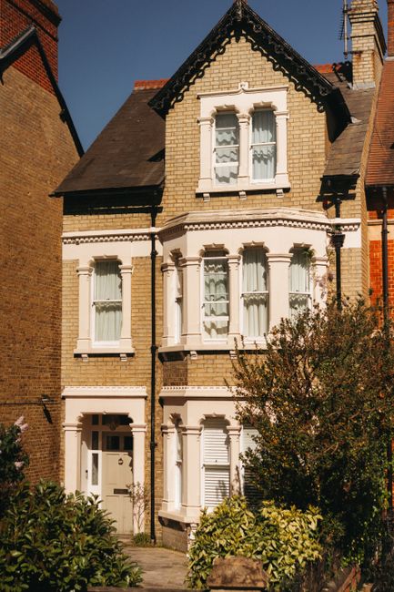A row of Victorian-style residential buildings constructed from grey stone with pitched roofs and chimney stacks, set along a tree-lined street in Kentish Town. The front gardens are neatly maintained